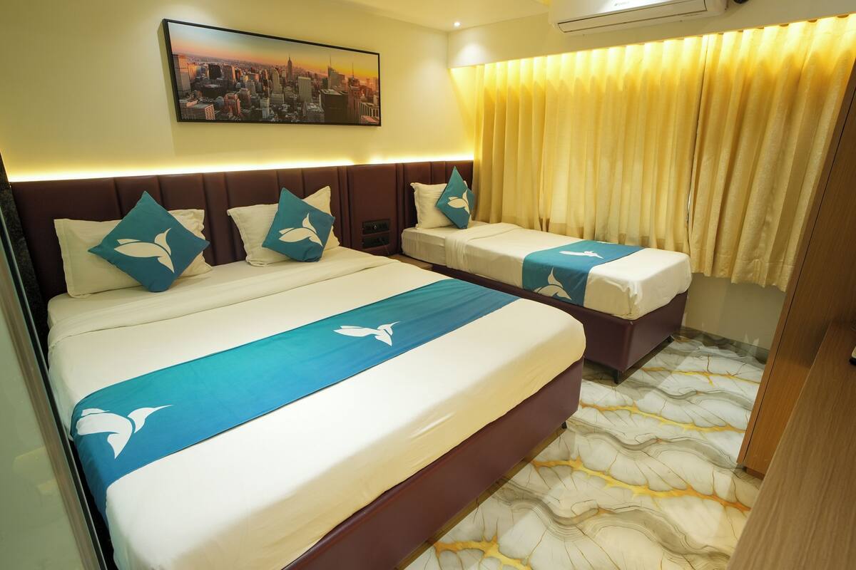 Een hotelkamer met een groot bed, twee kleinere bedden, een stadslandschap schilderij en een vloer met een patroon.