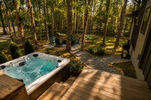 A hot tub in a forested backyard with a deck, outdoor furniture, and a barbecue grill.