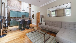 A modern kitchen with teal cabinets, a dining area with two chairs, a sofa, and a large mirror. - Charming One-Bed Oasis in Hammersmith (London)