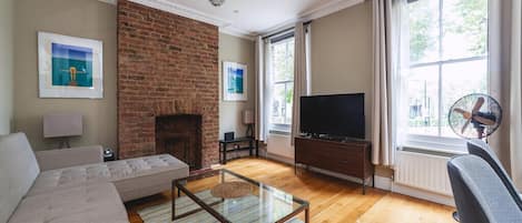 A living room with a brick fireplace, a sofa, a glass coffee table, a TV, and a large window with curtains.