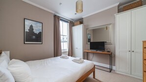 A bedroom with a bed, a television on a stand, a wardrobe, and a window with curtains. - Charming One-Bed Oasis in Hammersmith (London)