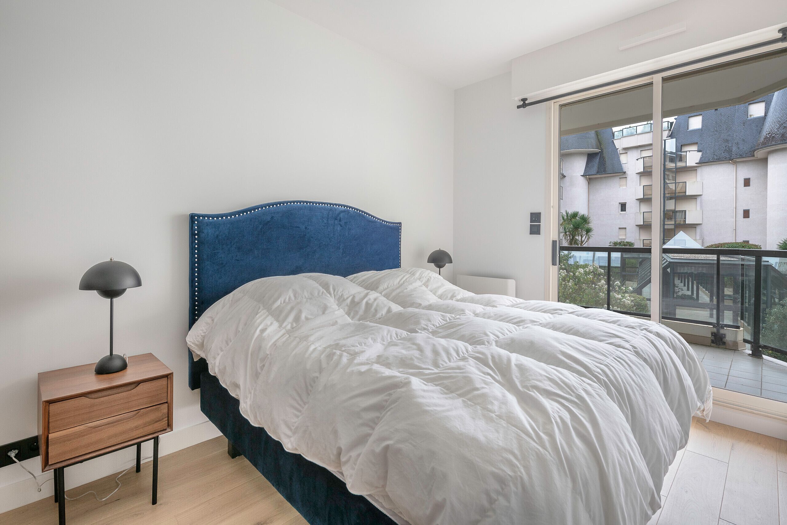 Una camera da letto con un letto grande, un comodino con una lampada e vista sugli edifici da un balcone.