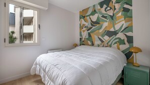 Une chambre avec un grand lit, une table de chevet verte, une lampe fixée au mur et une fenêtre donnant sur un immeuble.