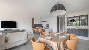 Une cuisine moderne dotée d’un îlot central, un coin repas avec une table dressée pour six personnes, et un espace salon avec un canapé et une télévision.