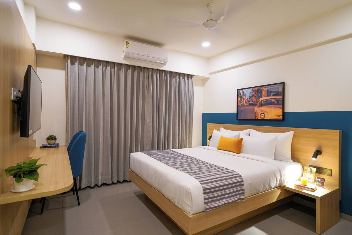 A hotel room with a bed, a desk with a chair, a TV, an air conditioner, and a painting on the wall.