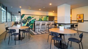 A modern indoor space with round tables and black chairs, featuring a checkered floor pattern and a large green and yellow abstract wall art piece. - Olive Baner by Embassy Group (Pune)
