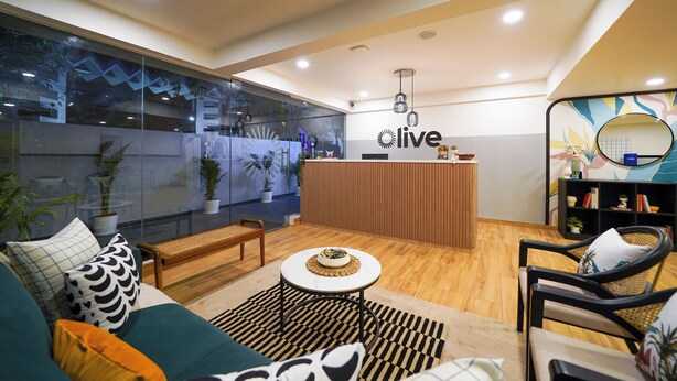 A modern living area with a reception desk labeled 'live', a sofa with patterned cushions, a coffee table, and a bookshelf with decorative items. - Olive Baner by Embassy Group (Pune)