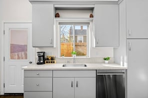 Una cocina moderna con armarios grises, lavavajillas de acero inoxidable, cafetera y ventana con vista a una valla de madera y edificios.