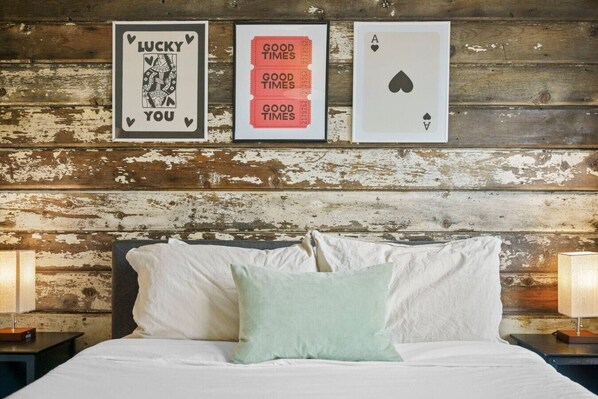 A bed with pillows, a wooden wall, and three framed items: a playing card, a card with 'GOOD TIMES' text, and a card with 'LUCKY YOU' text.