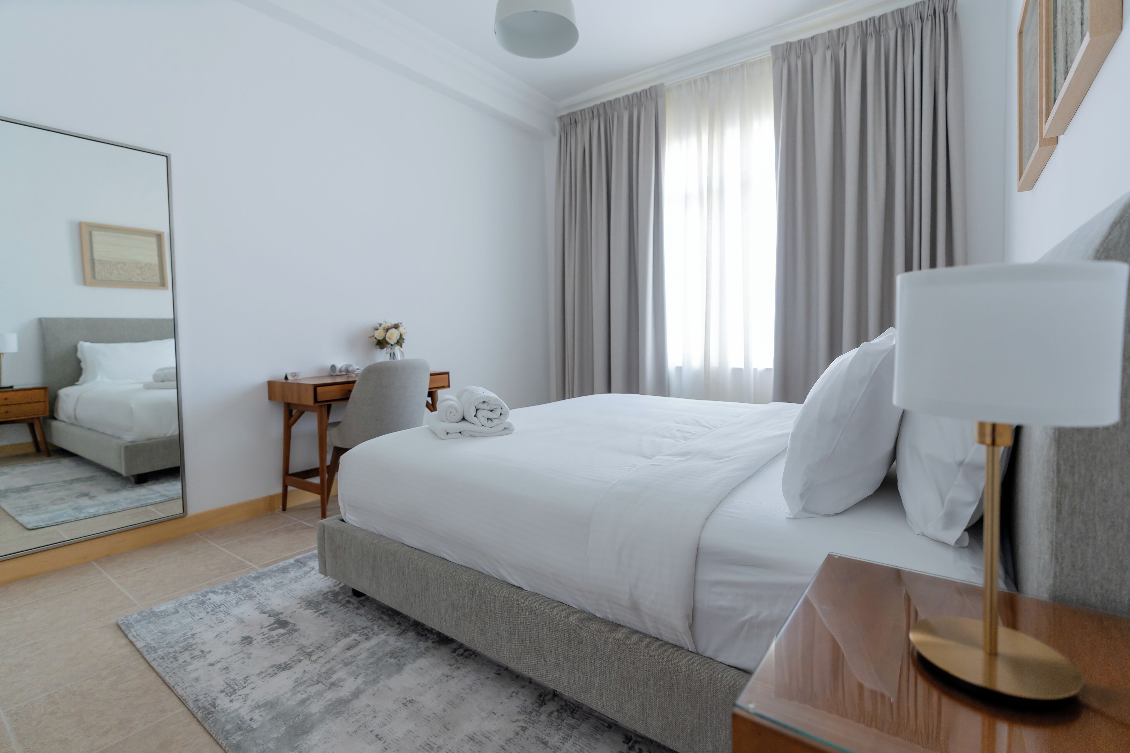 A hotel room with a bed, a desk, a chair, a mirror, and a window with curtains.