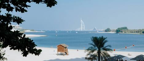 A beach with a wooden hut, palm trees, and a distant sail-shaped building.
