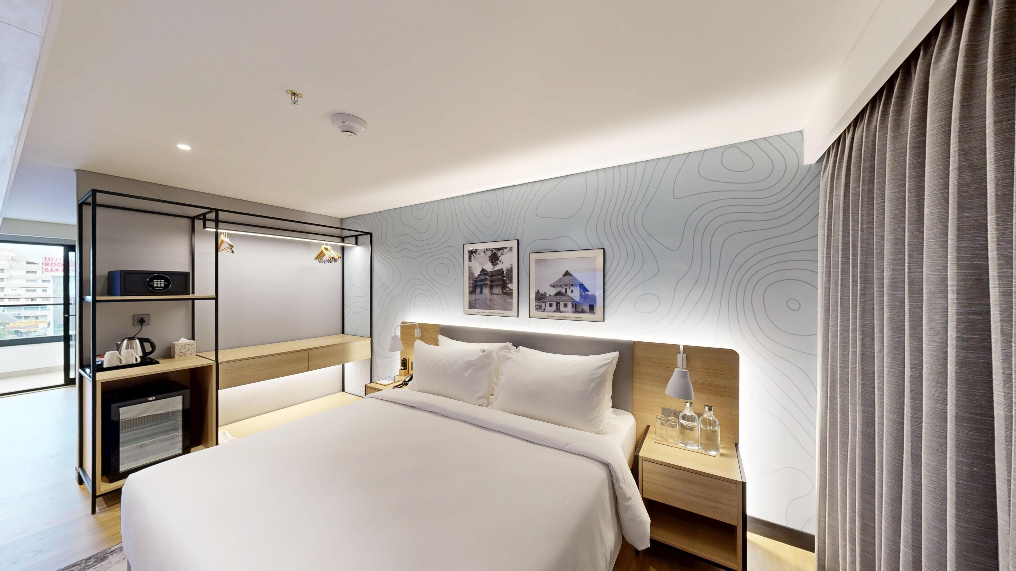 Una habitación de hotel moderna con una cama grande, una mesita de noche con lámpara, un armario y un pequeño escritorio con hervidor.