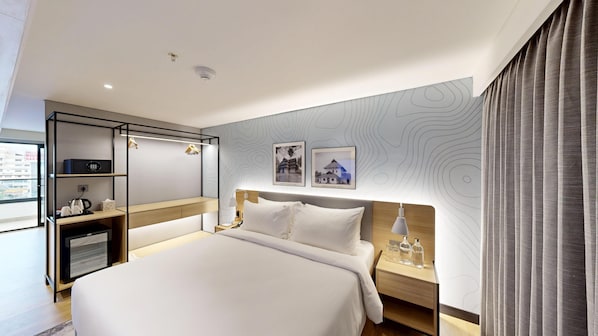 Quarto de hotel moderno com uma cama grande, uma mesa de cabeceira com abajur, um guarda-roupa e uma pequena escrivaninha com chaleira.