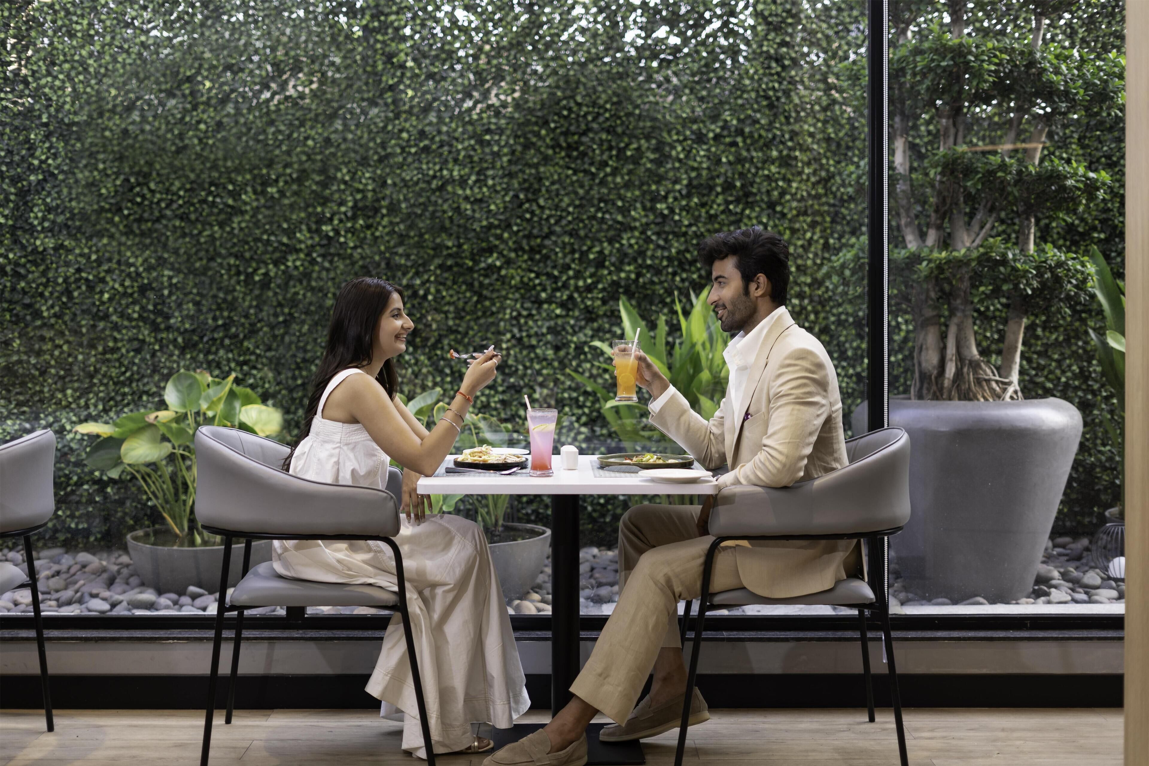 Two people seated at a table with a drink and food, surrounded by greenery and plants.