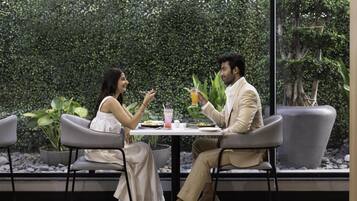 Two people seated at a table with a drink and food, surrounded by greenery and plants.