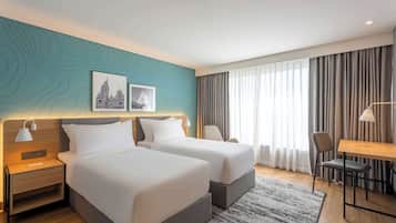 A hotel room with two beds, a desk, a chair, and a large window with curtains.