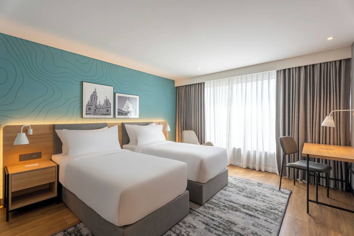 Une chambre d’hôtel avec deux lits, un bureau, une chaise, une table de chevet et une fenêtre avec des rideaux.