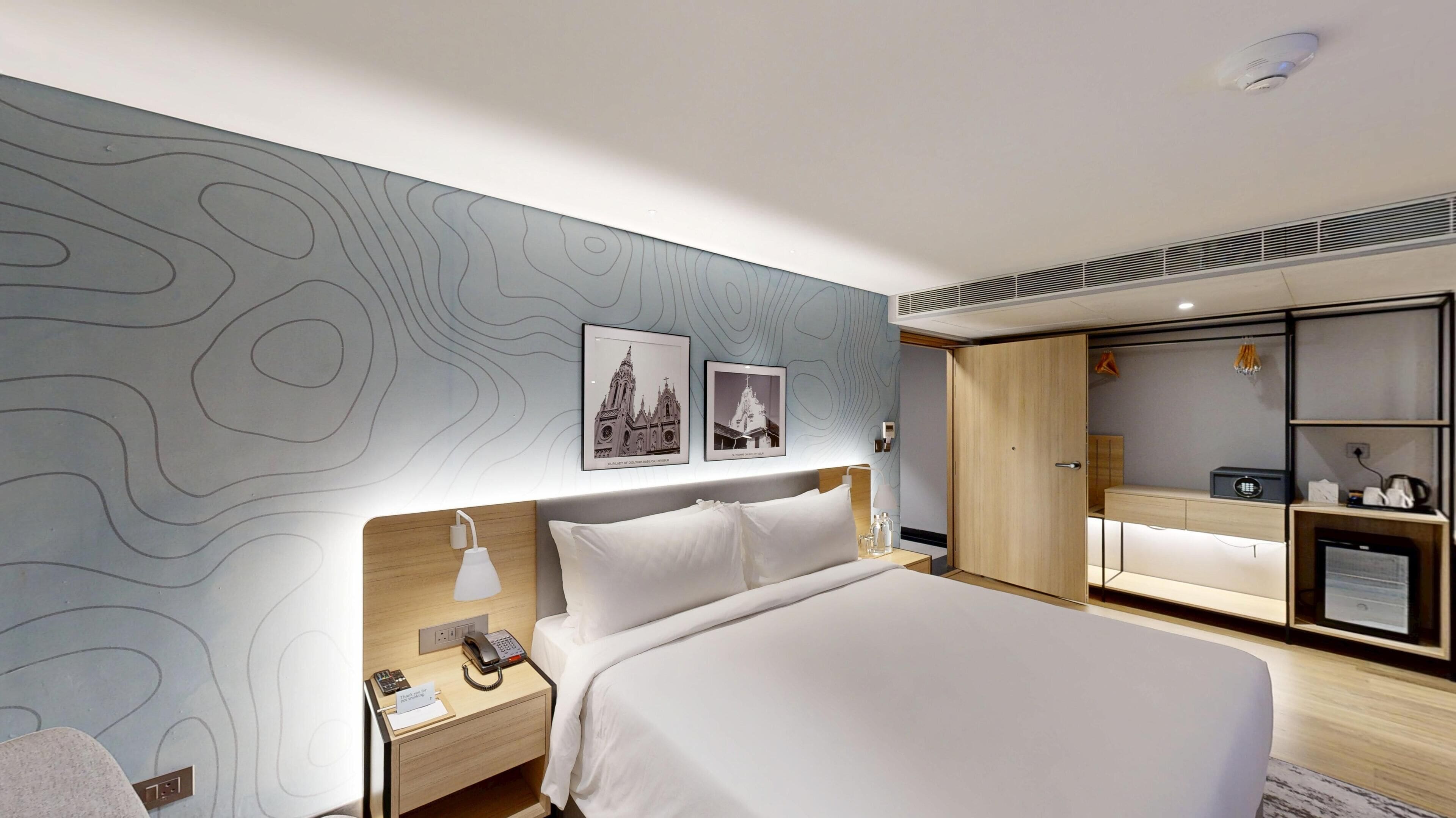 A modern hotel room with a large bed, a bedside table with a lamp, a nightstand with a phone and a notepad, a wardrobe, and a small desk with a radio and a kettle.