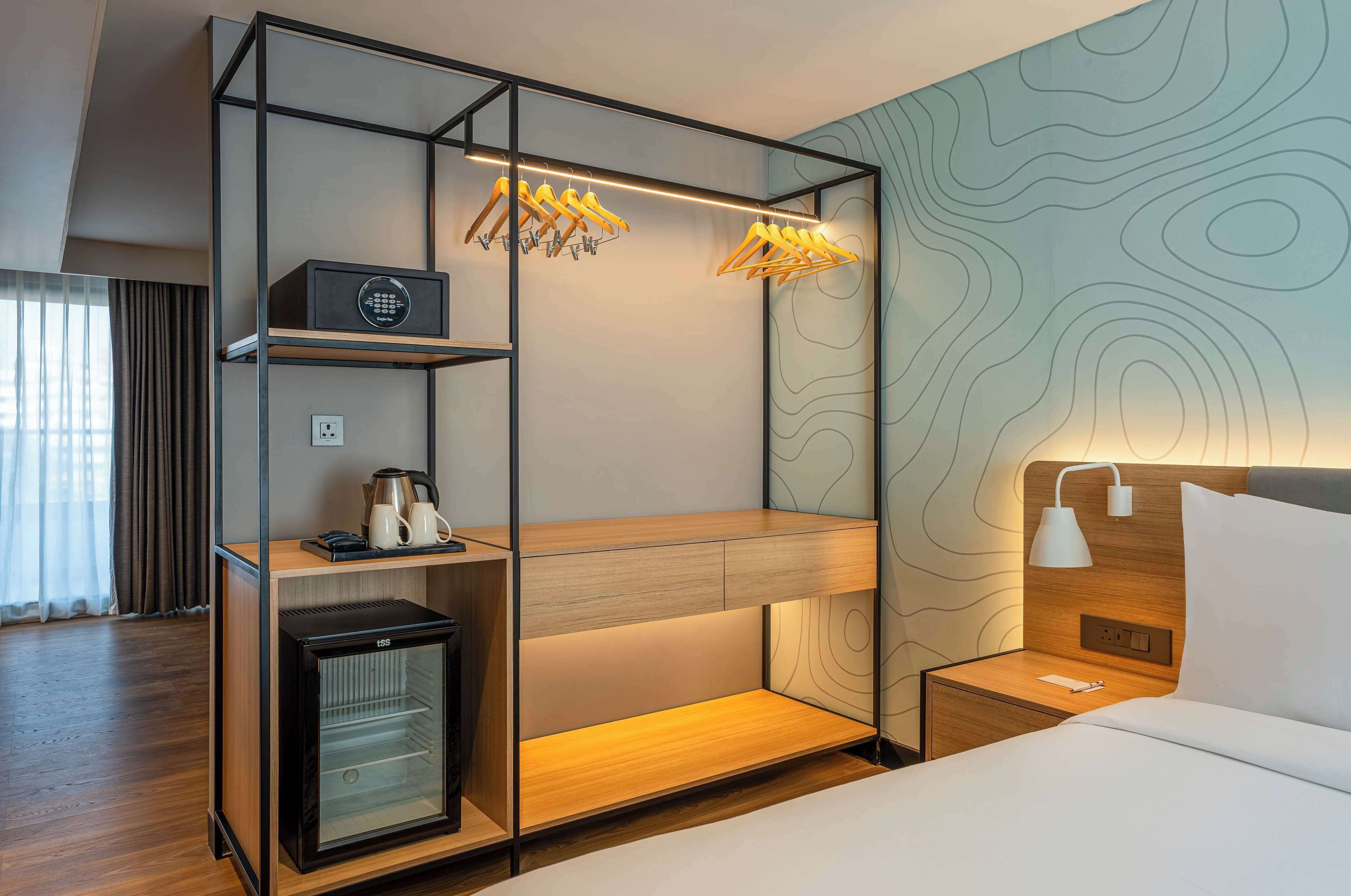 A compact hotel room with a bunk bed, a minibar, a small refrigerator, a bedside table with a lamp, and a wall with a topographic design.