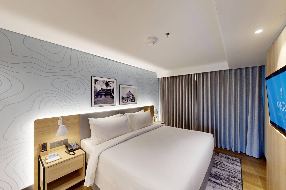 Une chambre d’hôtel moderne dotée d’un grand lit, d’une table de chevet avec une lampe, d’un écran de télévision affichant le logo « PARK INN & SUITES », et d’un mur orné d’un motif topographique.