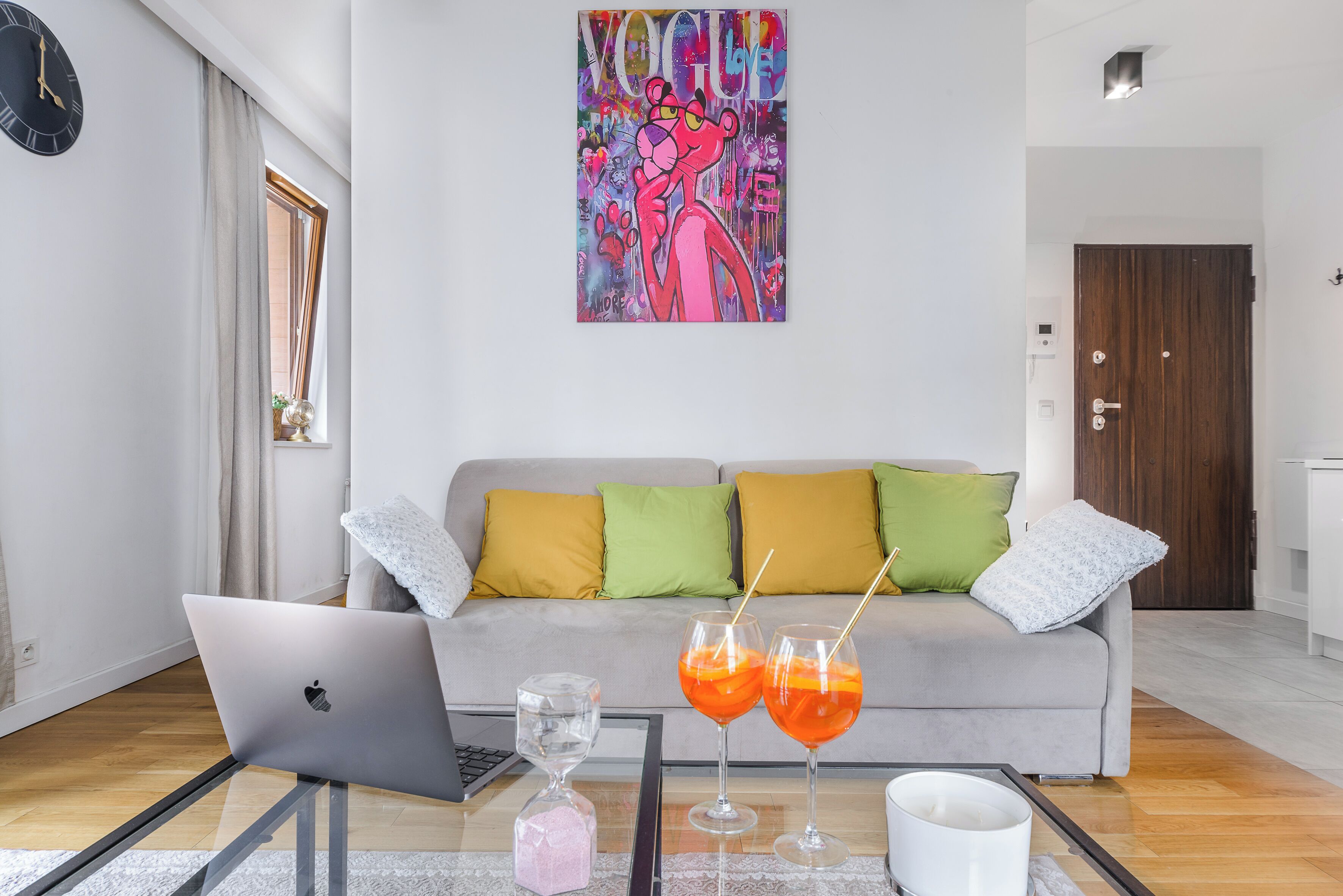 A modern living room with a grey sofa, colorful cushions, a laptop, and two drinks on a glass table.