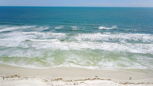 Ein Strand mit ankommenden Wellen, blauem Himmel und einigen Personen in der Ferne.