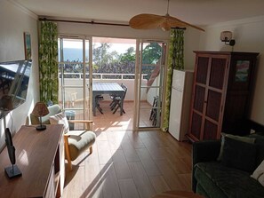A living room with a wooden table, a sofa, a TV, and a sliding glass door leading to a balcony.