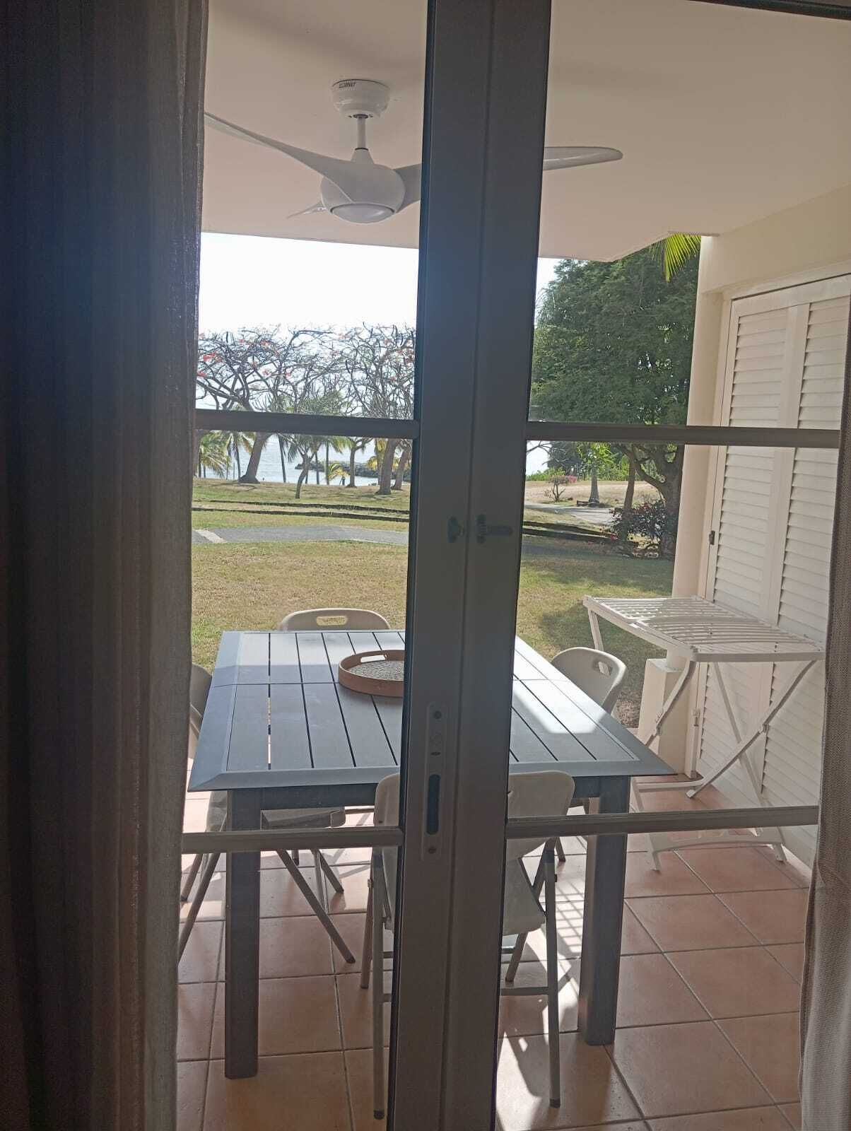 An open sliding door leading to an outdoor patio with a table and chairs, a ceiling fan, and a view of a grassy area with trees and a body of water.