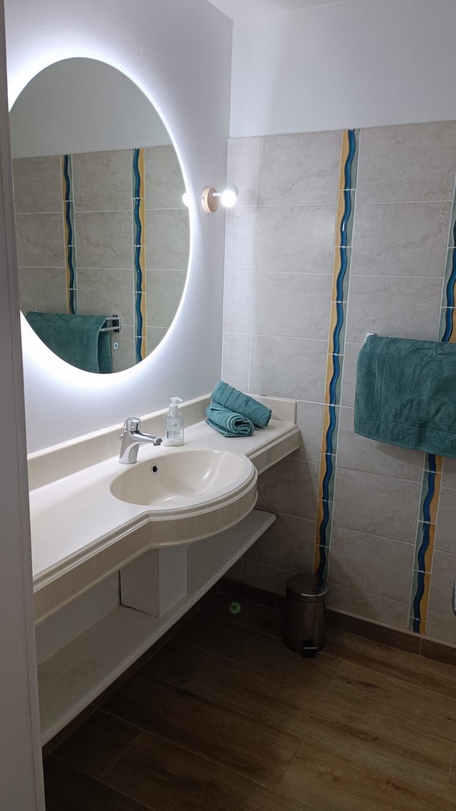 Un baño con un espejo redondo, un lavamanos, un perchero para toallas y un cubo de basura.