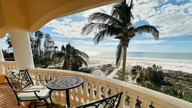 A balcony with two chairs and a table, overlooking a beach with palm trees and a clear sky. - St Pete Beach Don Cesar Beachfront Hot Tub (St Pete Beach)