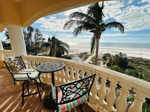 A balcony with two chairs and a table, overlooking a beach with palm trees and a clear sky.