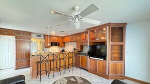A modern kitchen with wooden cabinets, a central island with bar stools, a ceiling fan, and a large flat-screen TV.