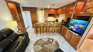 A modern kitchen with wooden cabinets, a central island with stools, a flat-screen TV, and a leather sofa in the living area.
