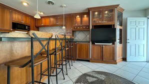 A kitchen with wooden cabinets, a bar area with stools, a microwave, and a flat-screen TV.
