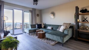 A modern living room with a green sectional sofa, two round side tables, a wooden coffee table, and a large potted plant. - Luxe Appartement Vista Maris Somaris - Havenweg 8-50 Sint-annaland Oosterschelde (Sint-Annaland)