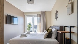 A modern hotel room with a large bed, a flat-screen TV mounted on the wall, a wooden nightstand, and a view of the city through the window. - Luxe Appartement Vista Maris Somaris - Havenweg 8-50 Sint-annaland Oosterschelde (Sint-Annaland)