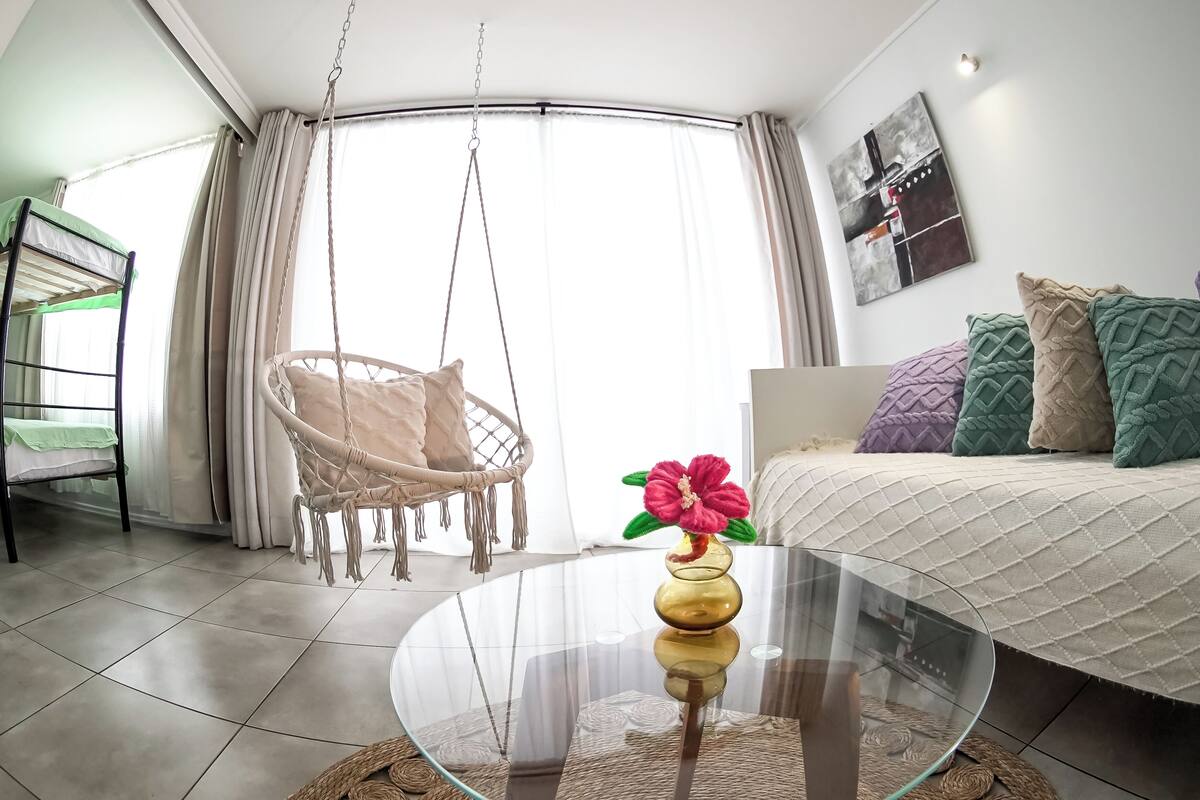 A modern living room with a glass coffee table, a hanging chair, a bed with patterned pillows, and a large window with curtains.