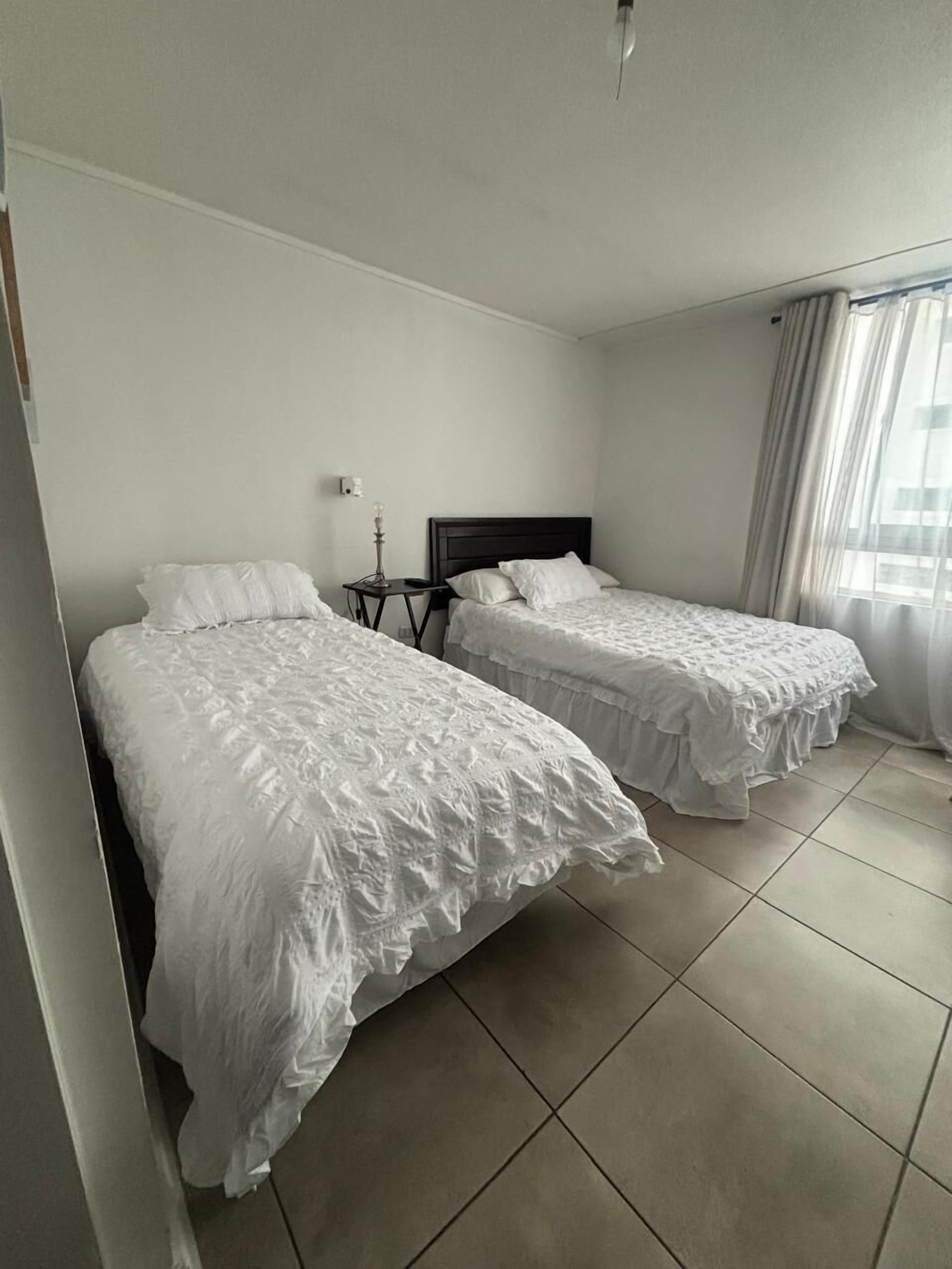 A hotel room with two beds, a nightstand, a lamp, and a window with curtains.