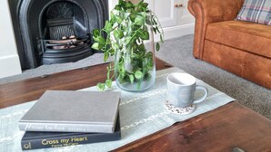 Ein Wohnzimmer mit Kamin, einem braunen Sofa, einem Couchtisch mit Büchern und einer Pflanze, sowie einer weißen Tür im Hintergrund.