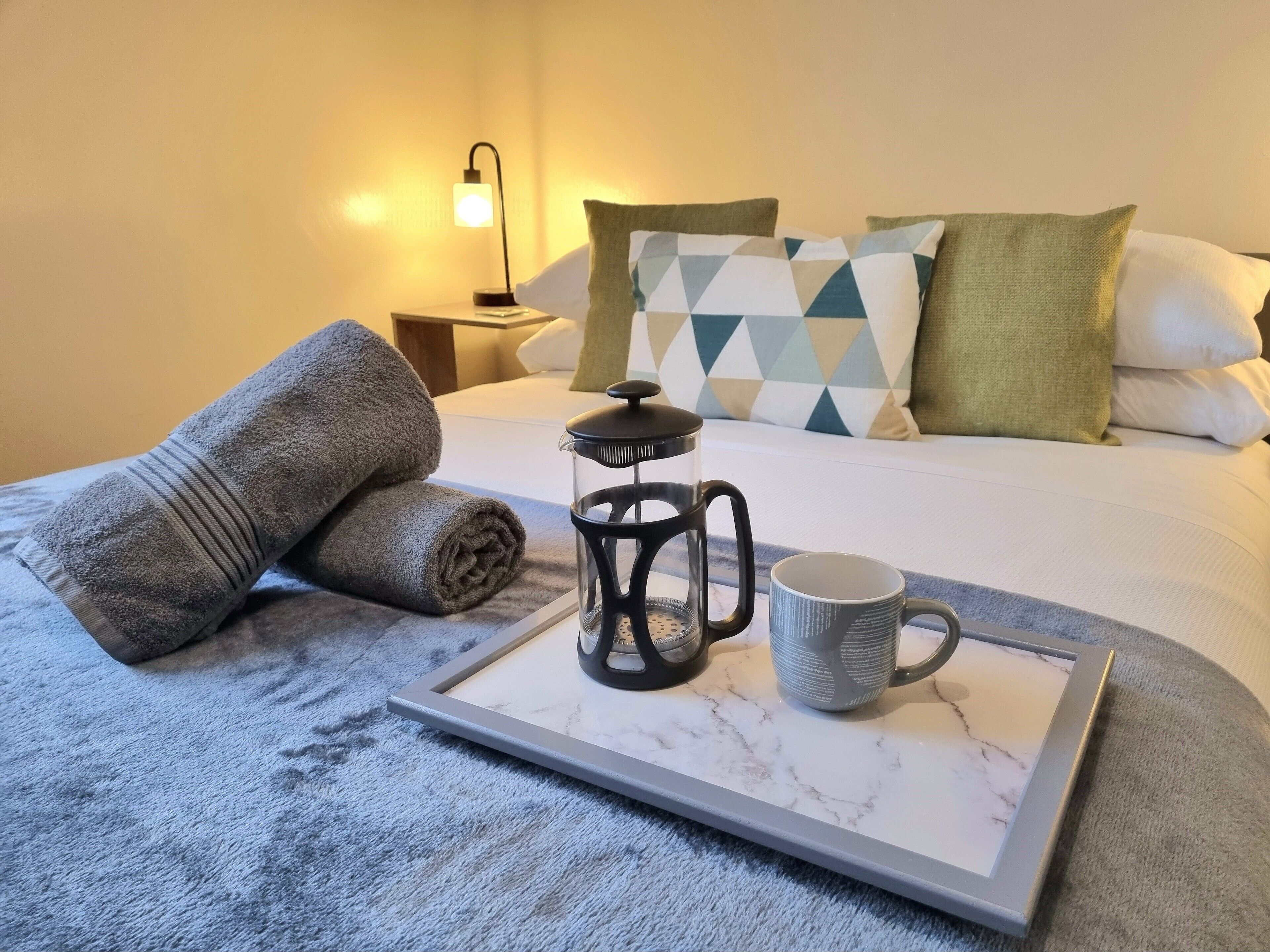 Una cama bien hecha con una toalla enrollada, una cafetera y una taza sobre una bandeja.