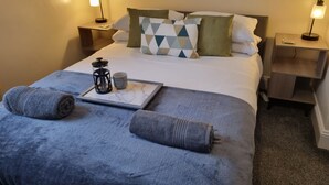 Ein ordentlich bezogenes Bett mit blauer Decke, zwei Kissen, einem Tablett mit Kaffeemaschine und Tasse sowie zwei Nachttischlampen.
