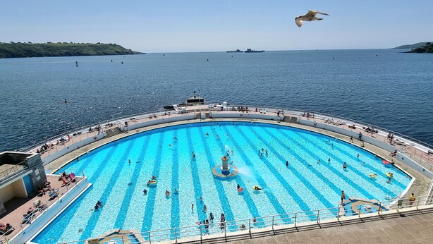 Ein großer Außenpool mit klarem, blauem Wasser, umgeben von einer Sonnenterrasse mit Sitzgelegenheiten und Blick auf das Meer.