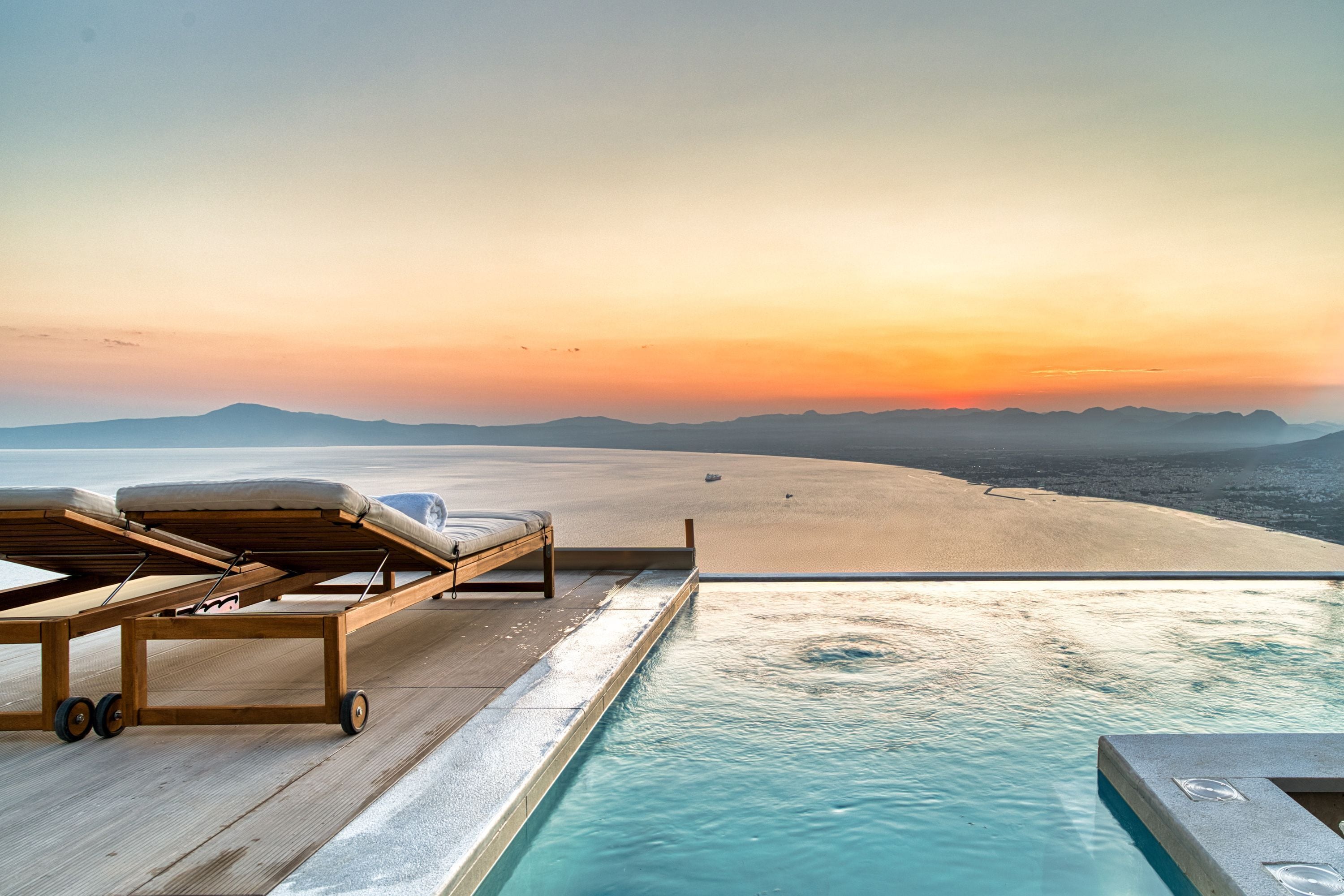 Un área junto a la piscina con vista a una playa al atardecer, una silla de descanso y una terraza de madera.
