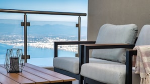 Un balcón moderno con vistas a la ciudad y montañas, que incluye una mesa de madera, dos sillas grises tapizadas y una baranda de metal.