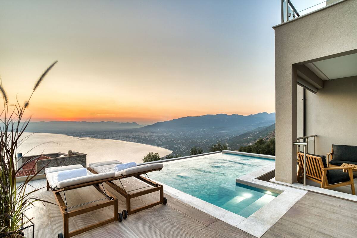 A rooftop infinity pool with lounge chairs, a view of the beach and mountains, and a modern architectural design.