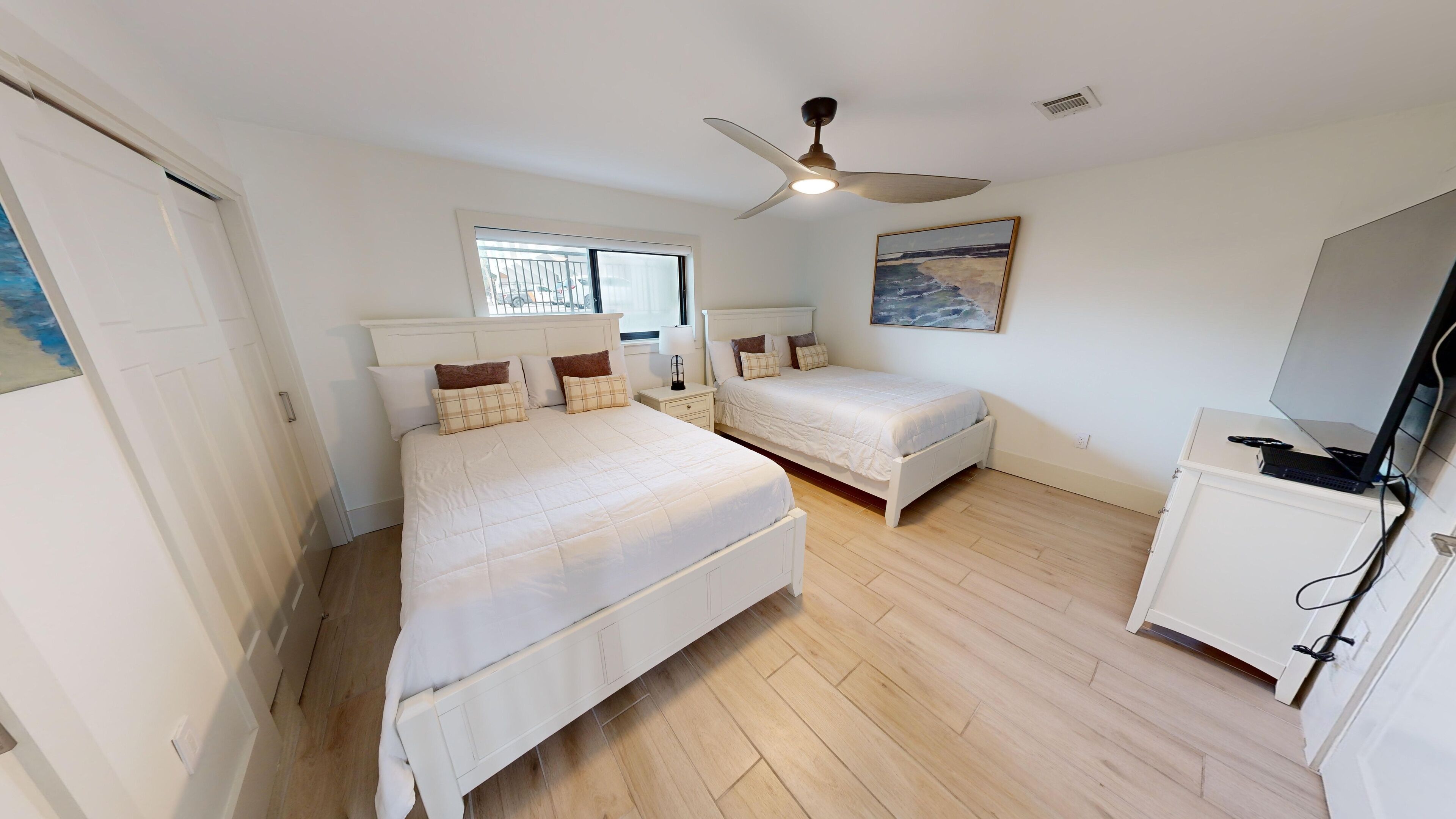 A bedroom with two beds, a ceiling fan, a window, a painting, and a desk with a computer.