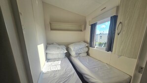 A small, compact room with two single beds, a window with curtains, and a wall-mounted shelf.