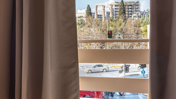 A room with a view of a cityscape, a bed with a blue cover, a glass table with a book and a cup, and a window with curtains. - Home of Petros | Near Plaka - Temple of Zeus View (Athens)