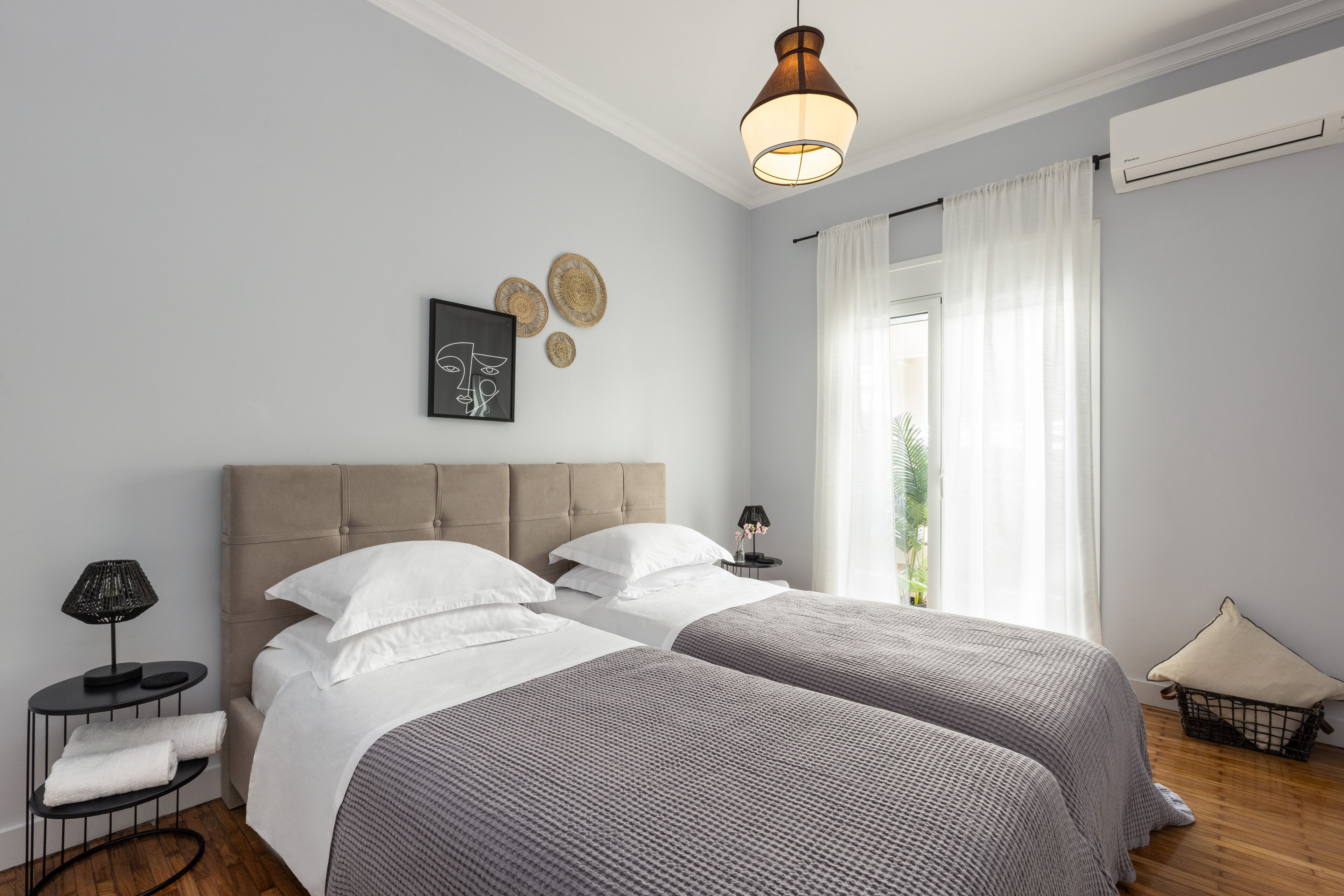 A modern bedroom with two beds, a bedside table with a lamp, a wall-mounted air conditioner, and a window with curtains.