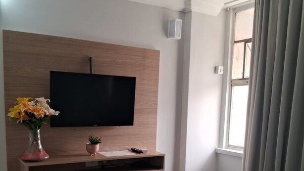 Une chambre d’hôtel avec un lit, un téléviseur fixé au mur, une petite étagère avec un vase de fleurs et une fenêtre avec des rideaux.
